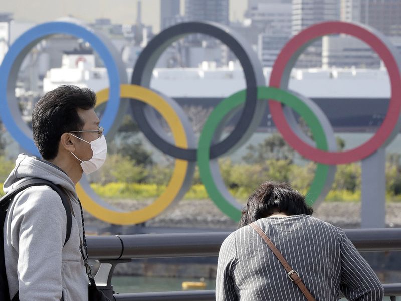 A man wearing a protective mask stands in front of Olympic rings in Tokyo