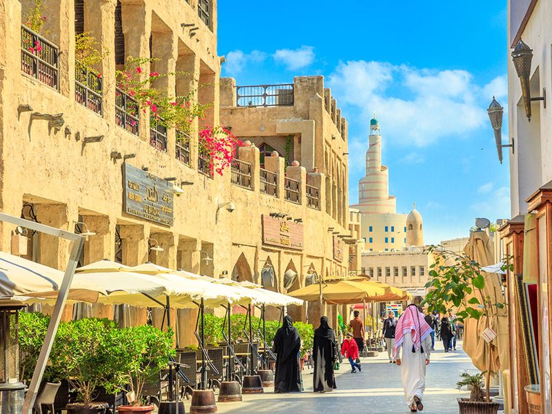Doha, Qatar - February 20, 2019: women and men in traditional Arab clothing walk along main street inside Souq Waqif old market with cafes and restaurants.Fanar Islamic Cultural Center in the distance.