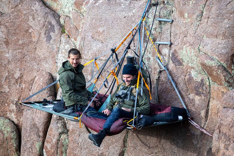 This image released by Nat Geo shows Bradley Cooper, left, and Bear Grylls on a paraledge hung off the edge of Pathfinder Canyon in Wyoming, in a scene from “Running Wild with Bear Grylls: The Challenge,