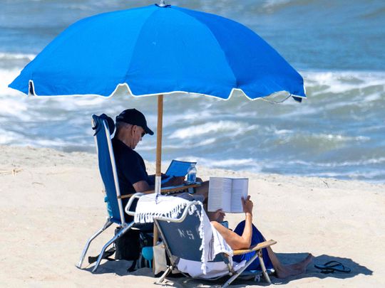 Beachgoers walk along the water as US President Joe Biden (L) and US First Lady Jill Biden sit under an umbrella in Rehoboth Beach, Delaware. 