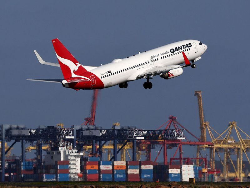 This picture taken on December 6, 2023 shows a Qantas Airways Boeing 737-800 passenger aircraft taking off at Sydney’s Kingsford Smith international airport in front of a container ship berthed at the Port Botany container terminal. (Photo by DAVID GRAY / AFP)