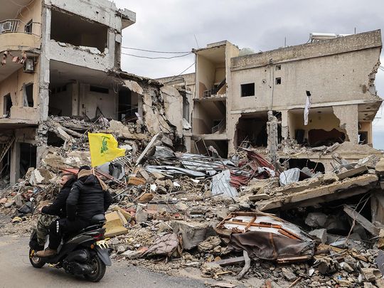 Men ride a scooter with a Hezbollah flag past destroyed buildings a